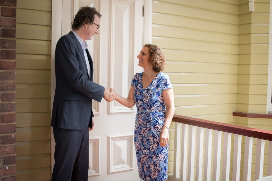 Tall man in suit shakes hands with smiling woman on steps of Queenslander house