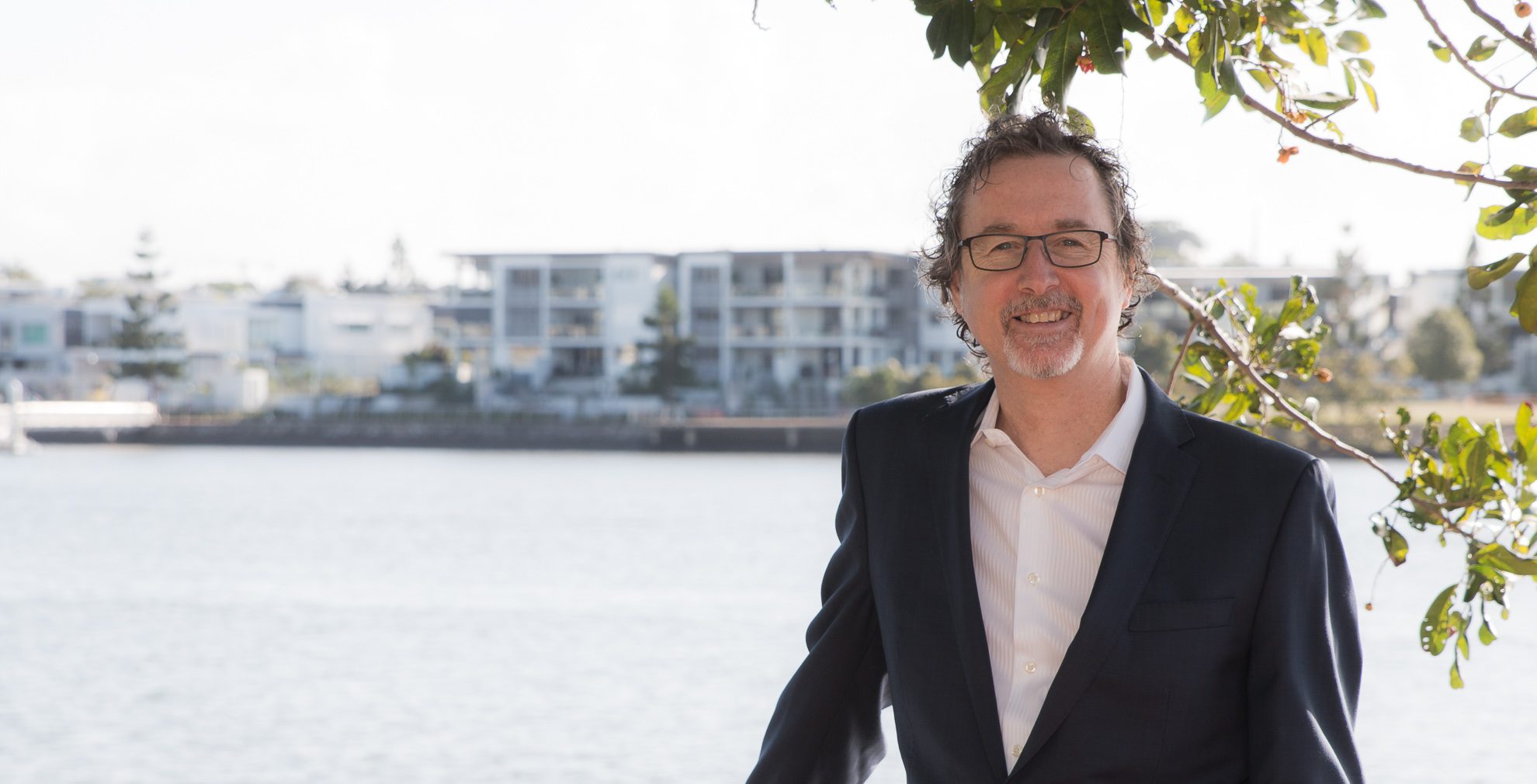 Man in 50s wearing navy jacket and white open neck shirt is smiling and standing casually in front of the Brisbane River. He is Derek Haas, mortgage broker, Brisbane. 