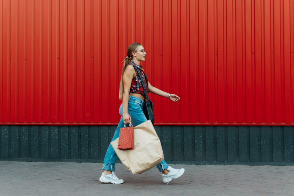 Happy girl with shopping bags walks past red wall. She is wearing blue jeans, white kicks and a checked top.